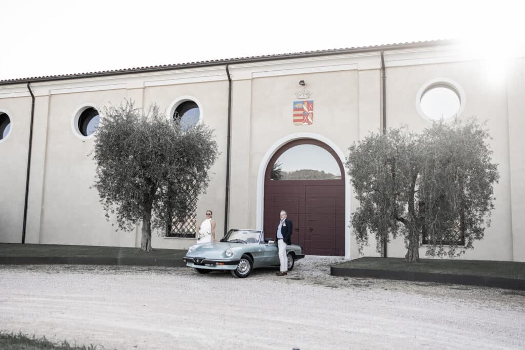 Ein Bräutigam steht an der Fahrerseite und die Braut an der Beifahrerseite eines alten offenen Cabrios der Marke Alpha Romeo. Im Hintergrund eine Mauer mit einem großen braunen Tor.