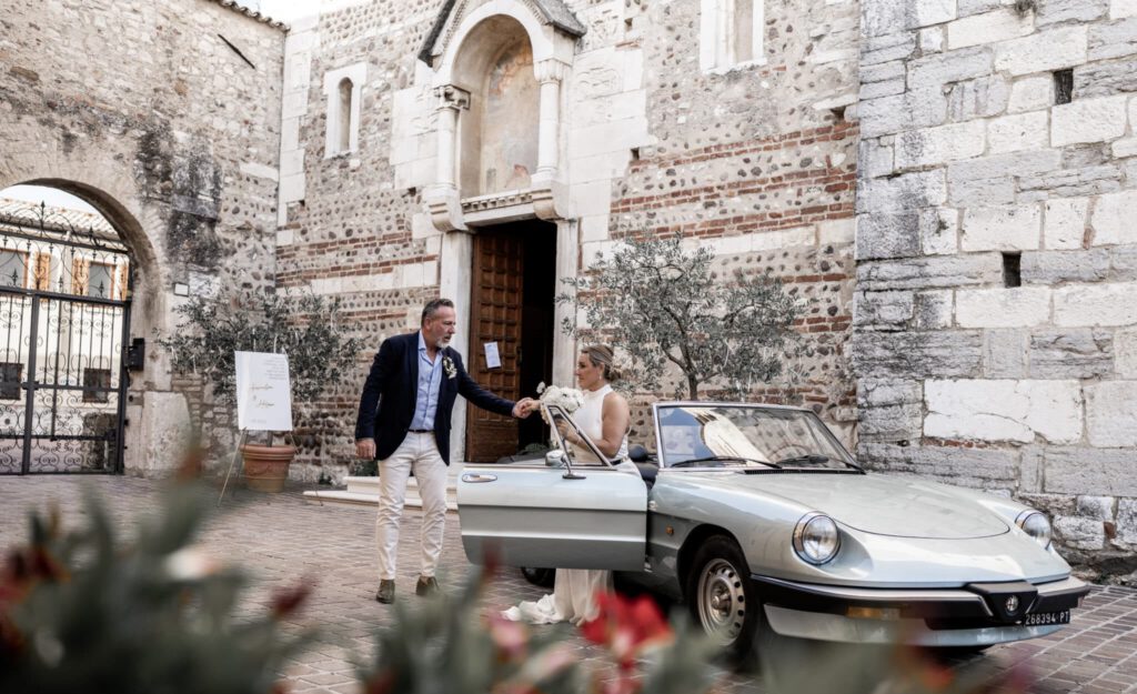 Ein Bräutigam hilft der Braut aus einem alten offenen Cabrio der Marke Alpha Romeo umgeben in einer steinigen Häuserkulisse.
