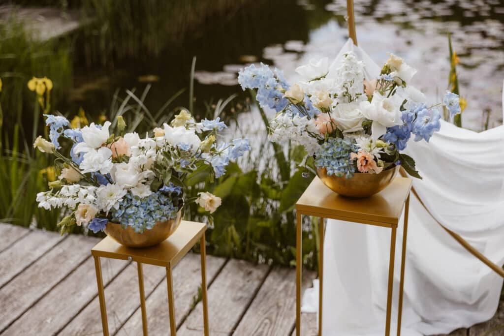 Zwei hellblau weiße Blumengestecke in matt goldenen Vasen auf goldenen Tischchen auf einem Holzboden ein weißen Tuch mit leichten Falten liegt im Hintergrund