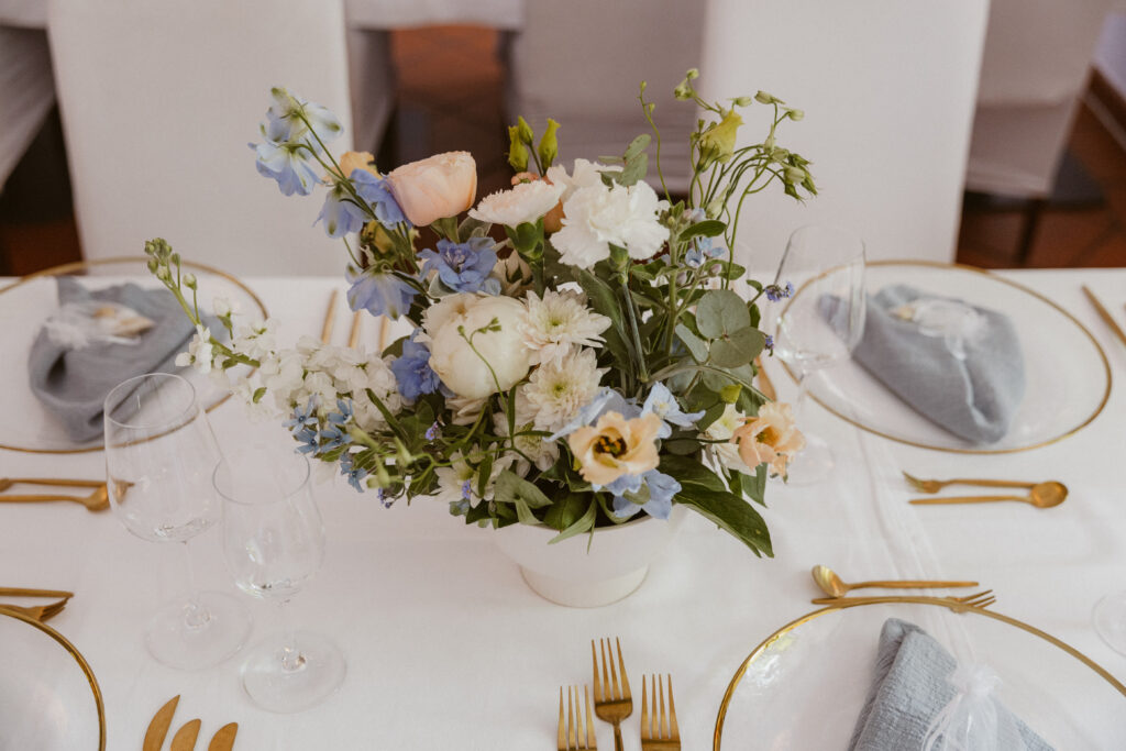 Ein gedeckter Tisch mit weißer Tischdecke. Goldenes Besteck neben goldumrandeten Tellern. In der Mitte steht ein blauweißer Blumenstrauß in einer weißen Vase