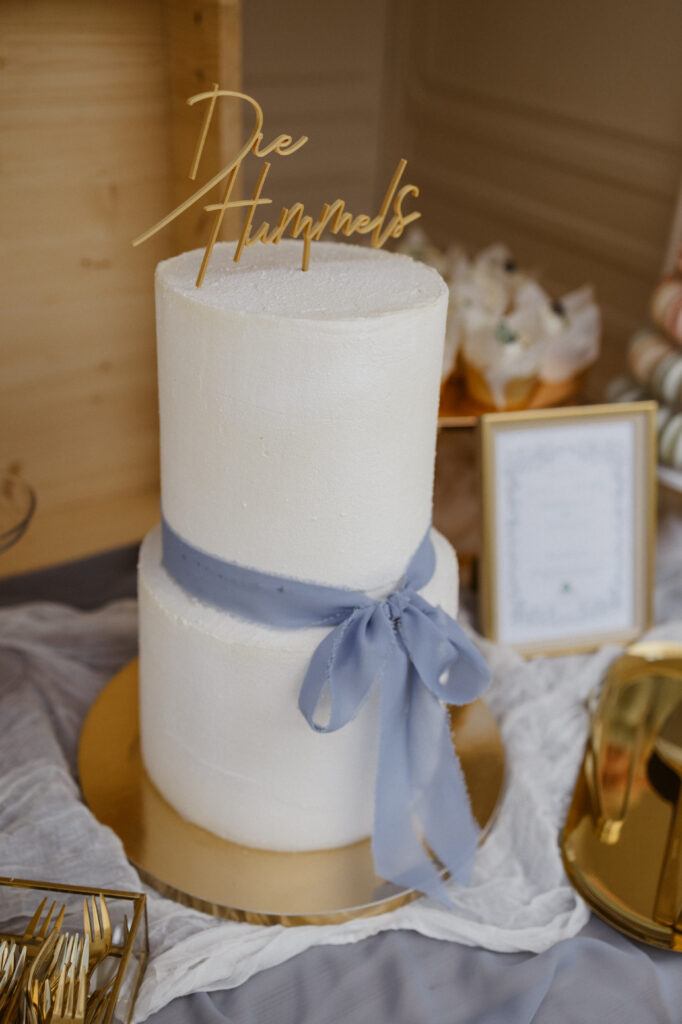 Eine zweistöckige weiße Hochzeitstorte, mit einer hellblauen Schleife geschmückt. Mit einem goldenen Schriftzug oben auf der Spitze 'Die Hummels'