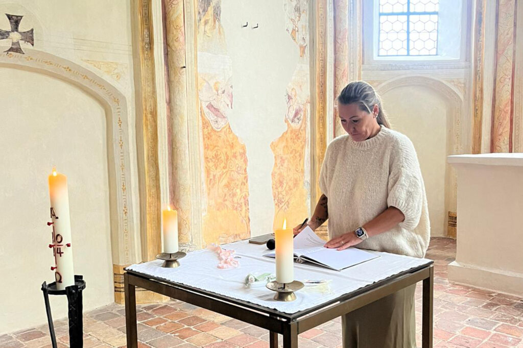 Die Traurednerrin steht vor einem Aufgeschlagenen Buch das auf einem Tisch in einer Kirche steht. Es brennen verschiedene Kerzen