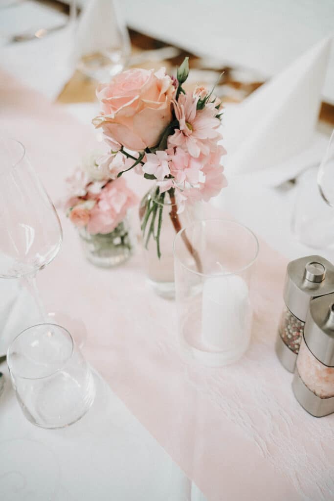 Eine kleine Glasvase mit einer großen rosa Rose daneben eine kleine Vase mit mehreren rosa Blumen, dezente Gläser auf der weißen Tischdecke.