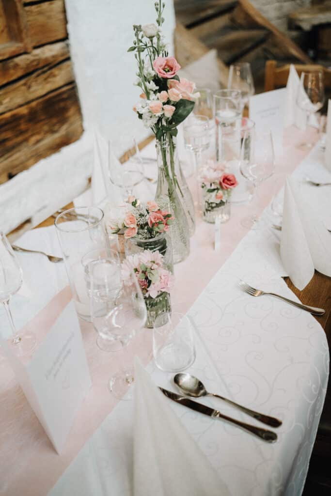 Ein gedeckter Hochzeitstisch mit rosa Banderole, rosa Rosen in Glasvasen und Gläser mit silbernem Besteck
