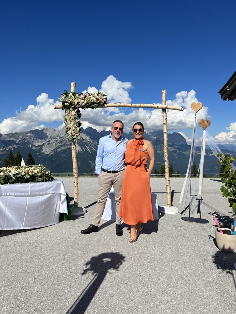Hochzeitsgäste mittlerem Alter stehen vor dem Traubogen. im Hintergrund ist ein Alpenpanorama zusehen