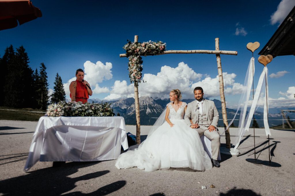 Eine Sprecherin in rotem Kleid traut ein Brautpaar und einem Traubogen aus weißen Birkenstämmen. Im Hintergrund sieht man die Alpen