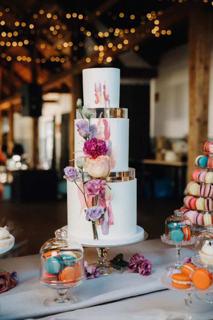 Eine dreistöckige weiße Hochzeitstorte, mit Rosen geschmückt. Daneben sind bunte Maceros