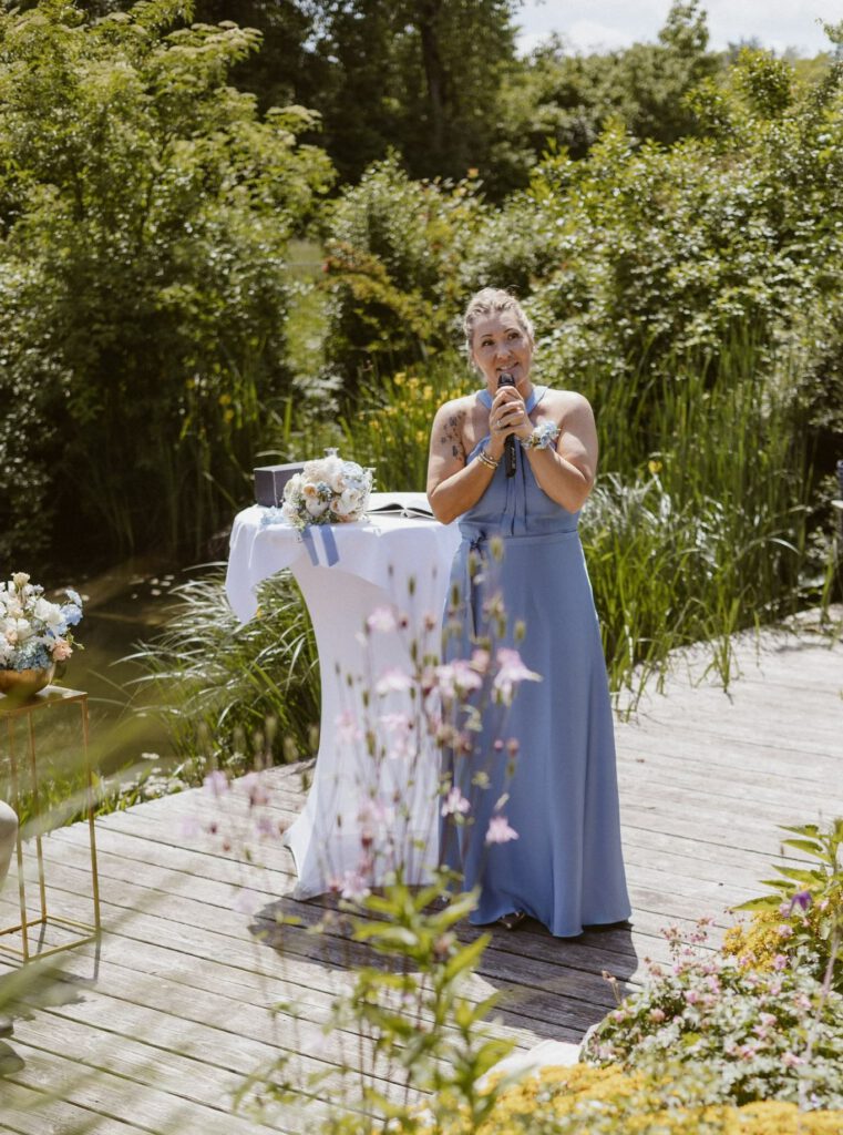 Eine hübsche Blond-, Braunhaarige Frau spricht in ein Mikrofon, sie hat ein langes lilafarbenes Fliederkleid an und steht auf meinem Steg, vor einem Teich mit leicht grünlichem Wasser. Neben ihr ist ein Stehtisch mit weißer Husse, darauf liegt der weißlich, rosa, hellblaue dezente Brautstrauß. Im Hintergrund befindet sich Schilf und andere Teichgräser.