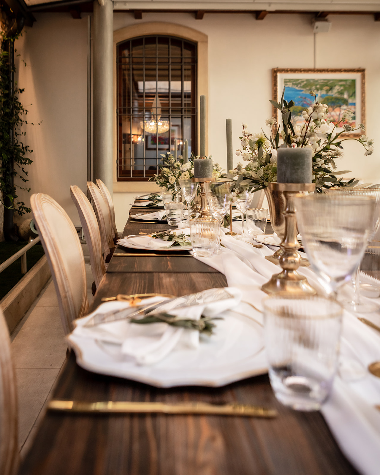 ein rustikaler Tisch gedeckt mit gold-weißen Tellern und goldenem Besteck. in der mitt liegt ein weißes stofftuch und goldene Kerzenständer schmücken die salbeigrünen Kerzen. Im hintergrund ist ein Fenster mit Eisenstäben die den Raum trennen