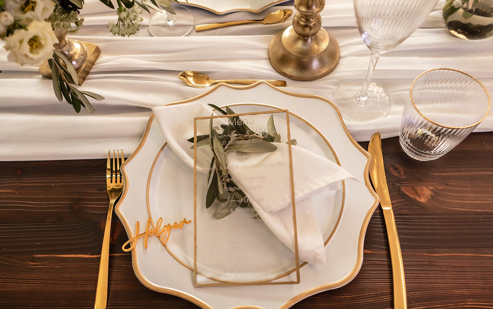 ein rustikaler Tisch gedeckt mit gold-weißen Tellern und goldenem Besteck. in der mitte liegt ein weißes stofftuch und goldene Kerzenständer schmücken die salbeigrünen Kerzen