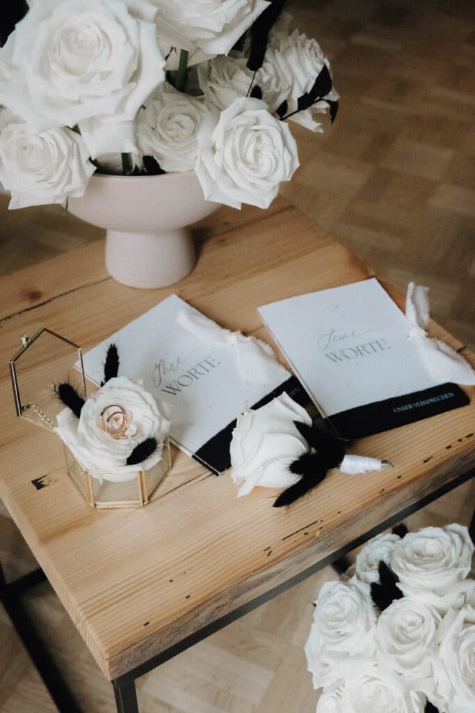 Weiße Rosen auf einem kleinem Holztisch, die Eheringe in einer Glasschatulle auf einer weißen Rose, zwei kleine Hefte mit der Aufschrift "The Wrote“ liegen im Vordergrund